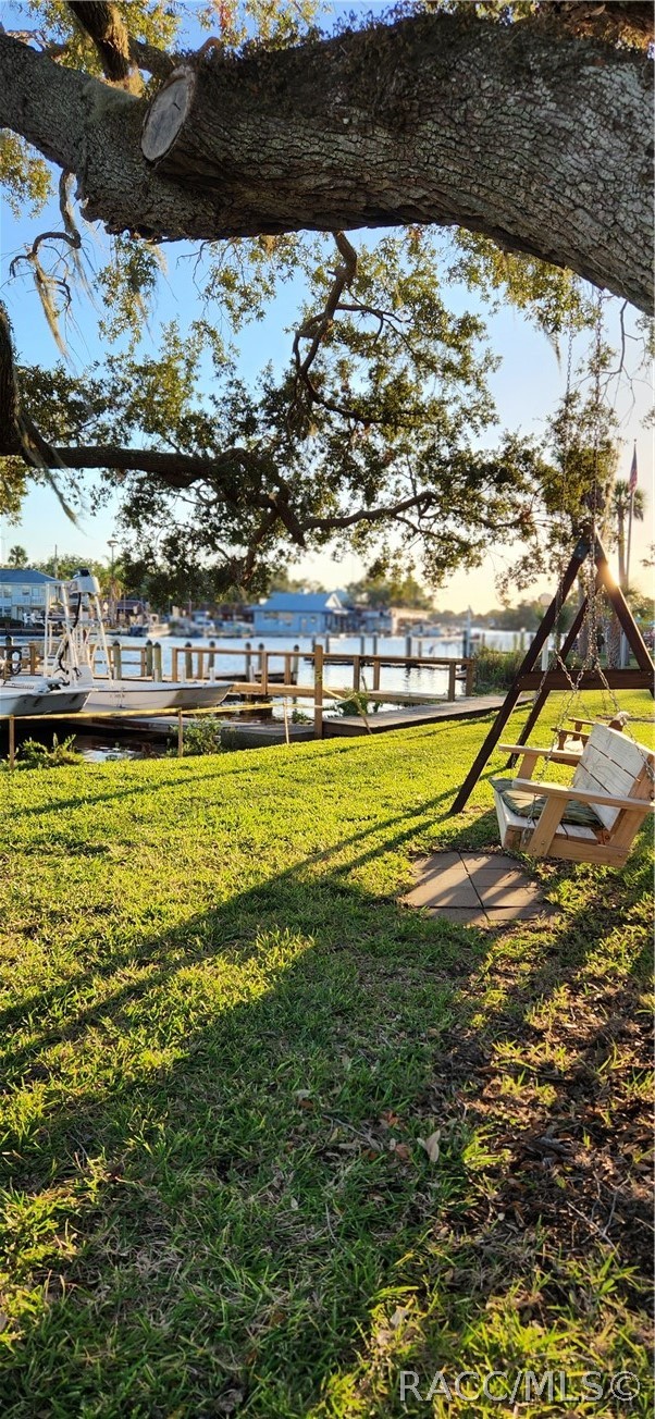 Waterfront yard with a wooden swing beneath a large oak tree, overlooking a dock and boats.