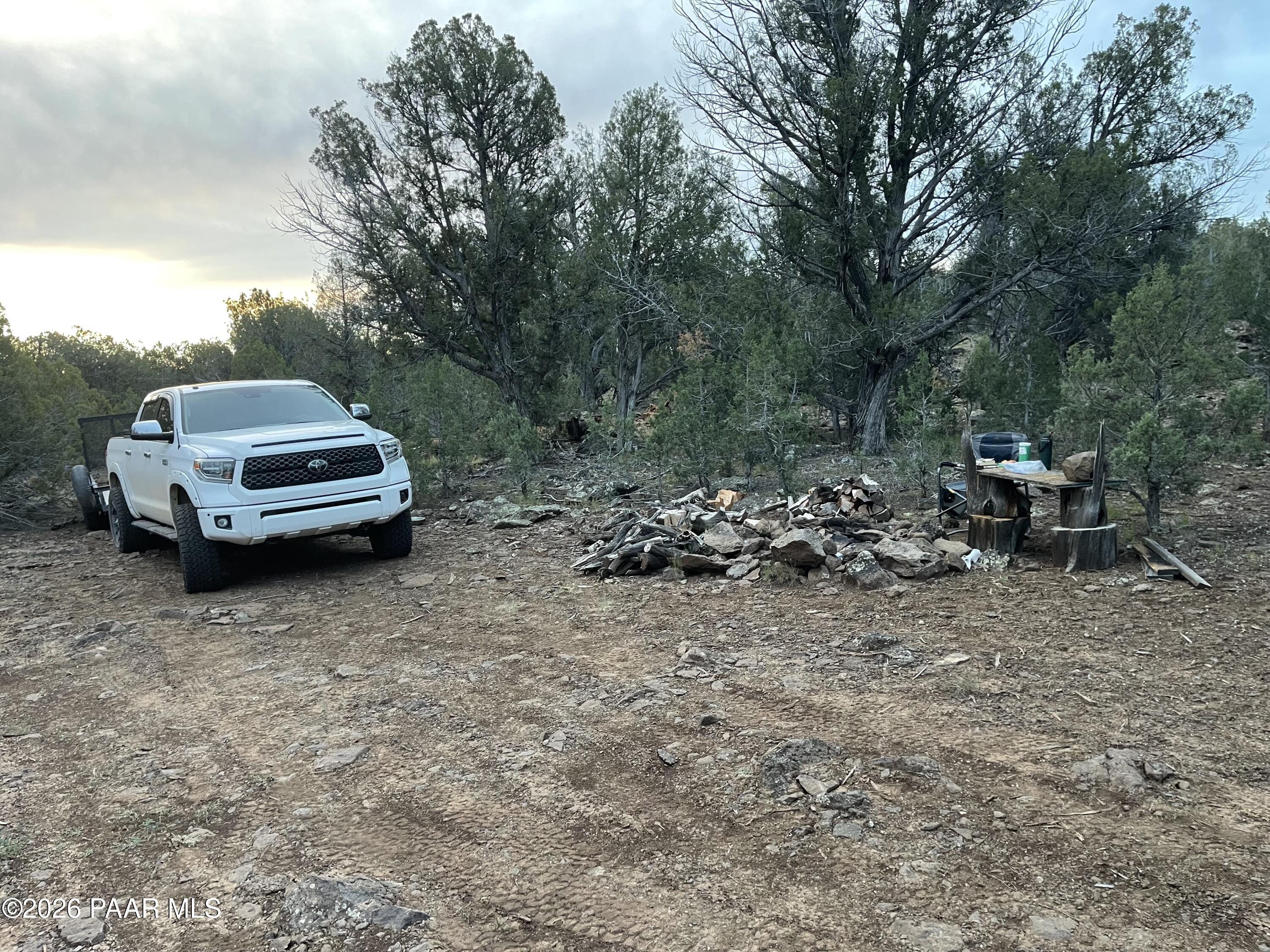 Wooded vacant lot with a rocky clearing, white pickup truck, firewood pile, and rustic outdoor seating.