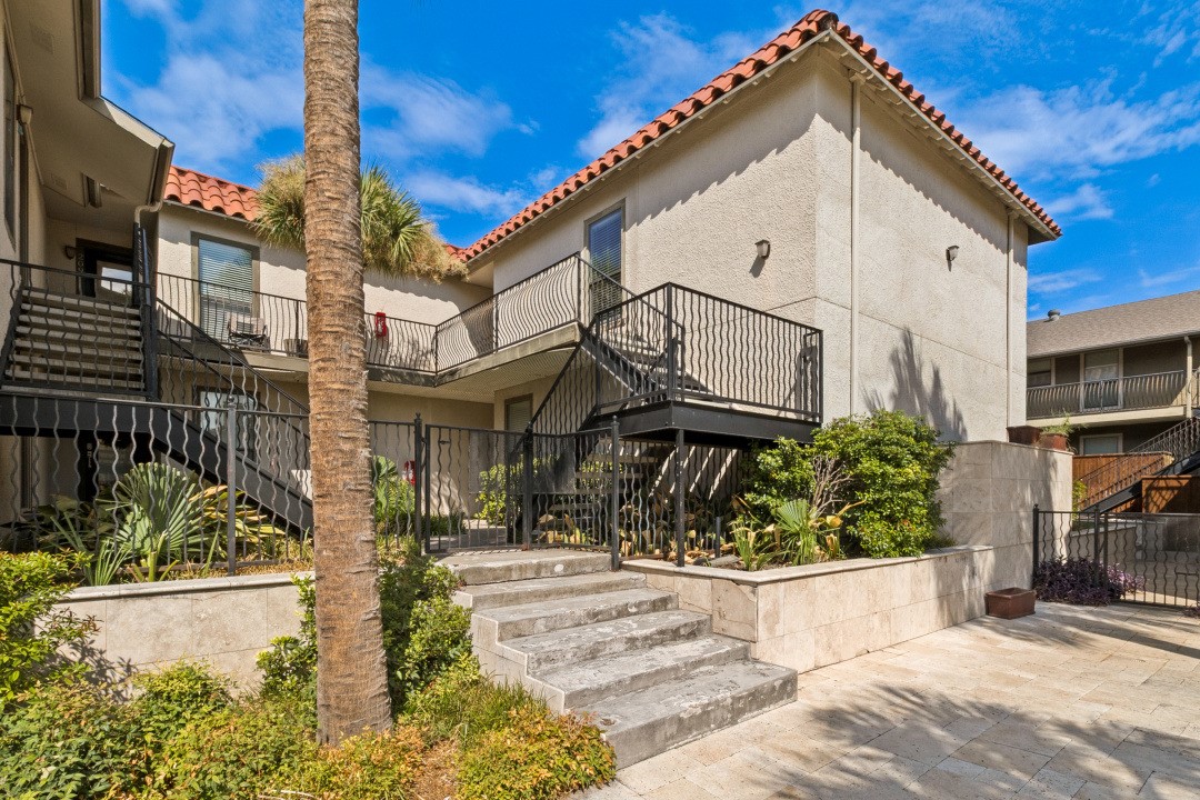 Residential building exterior with terracotta tile roof, metal balconies, landscaped beds, and stone courtyard steps.