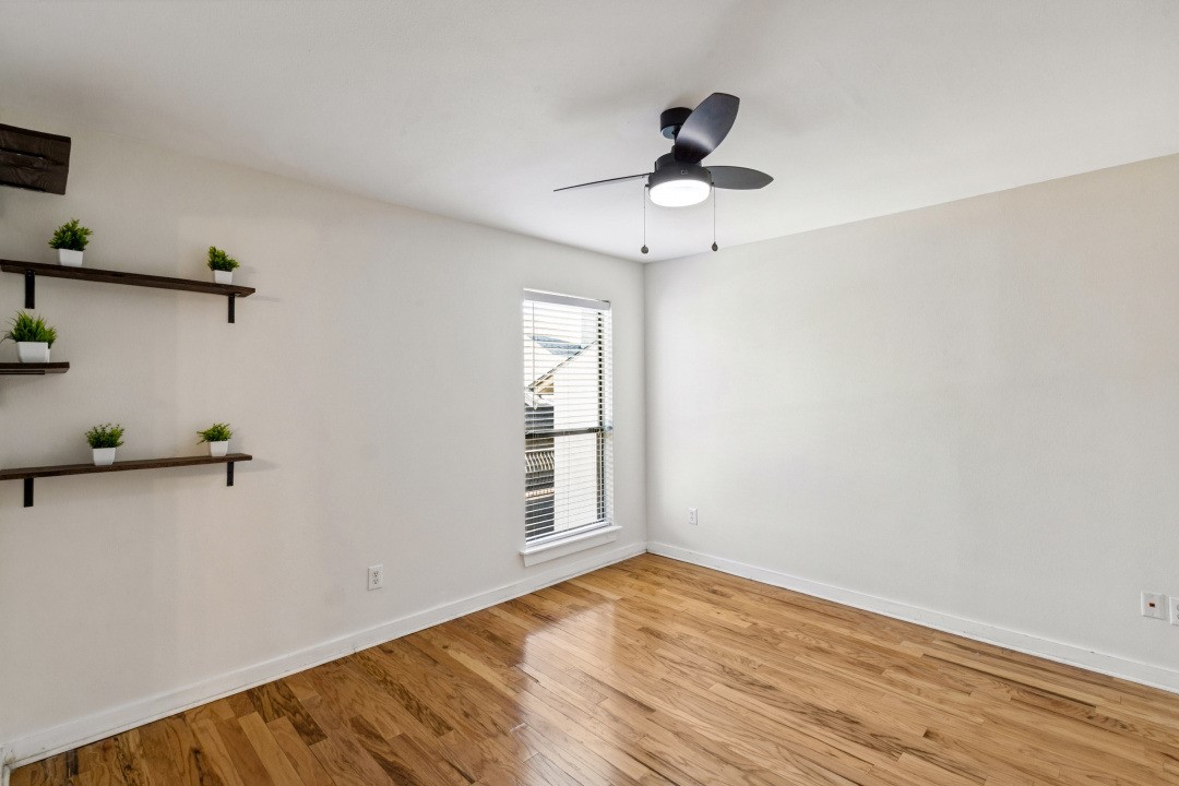 Empty room with polished hardwood floors, a ceiling fan, and floating shelves with potted plants.