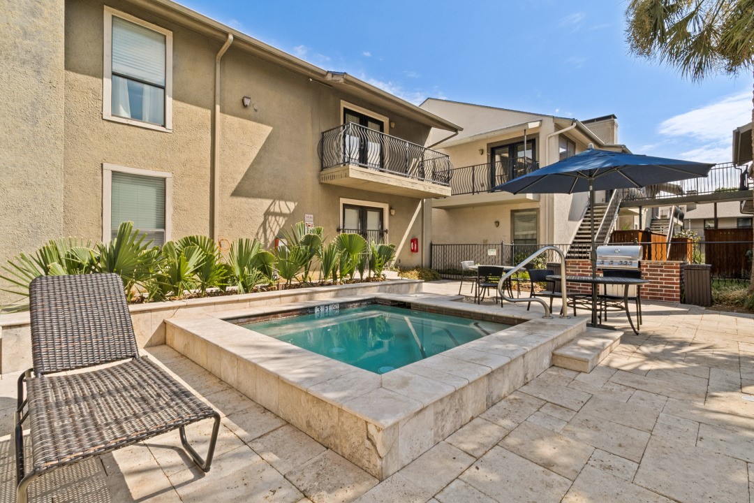 Courtyard pool beside a beige residential building, with stone paving, lounge chairs, and a blue umbrella.
