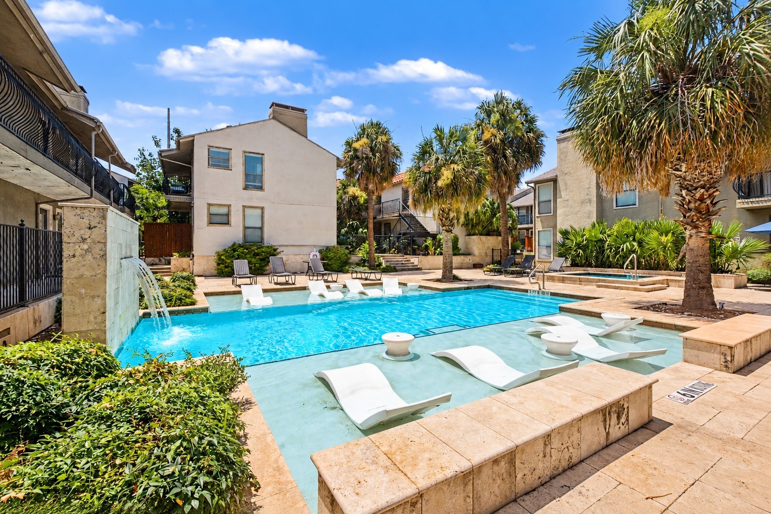 Residential complex pool with palm trees, a stone fountain, and white in-water lounge chairs.