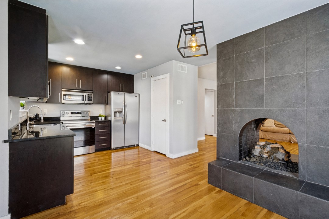 Open-plan kitchen with stainless steel appliances and hardwood floors beside a charcoal-tile fireplace.