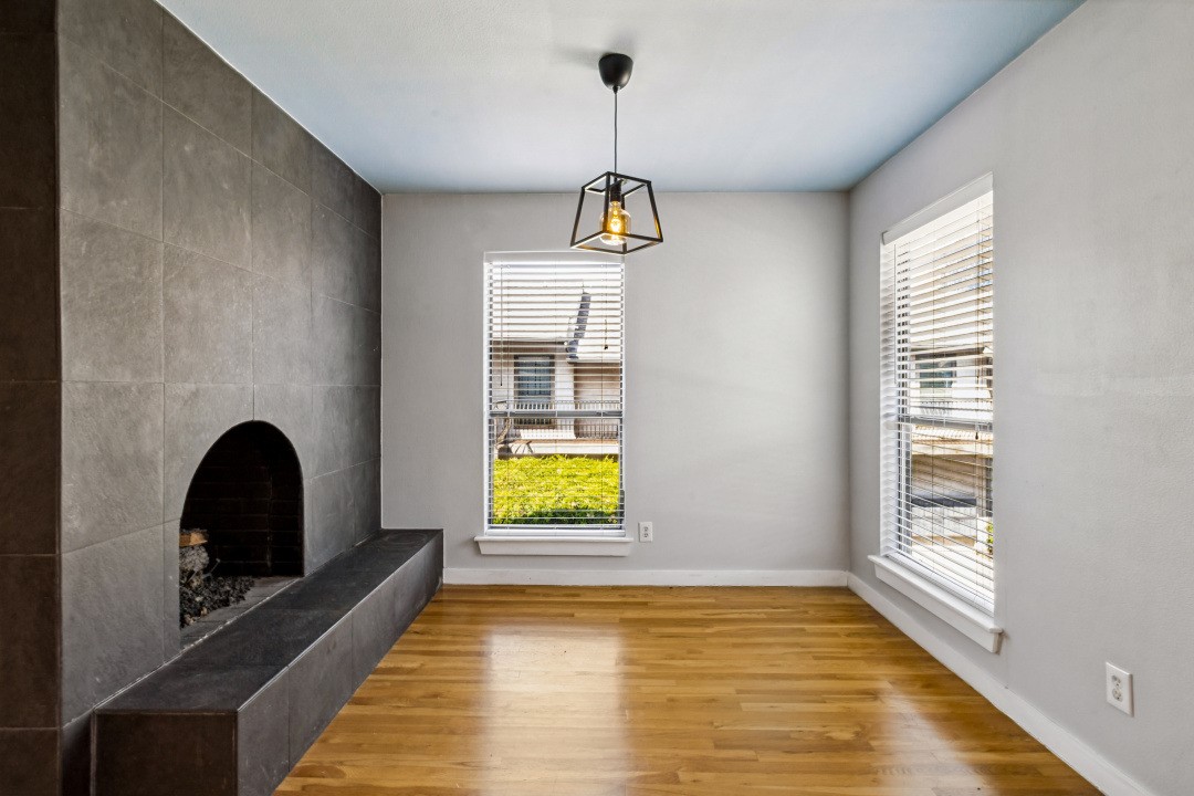 Empty room with a tiled fireplace, hardwood floors, and two windows with blinds.