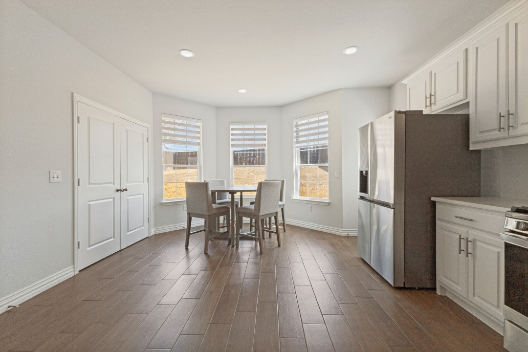 Kitchen dining area with white cabinetry, stainless refrigerator, and a table beside three windows.