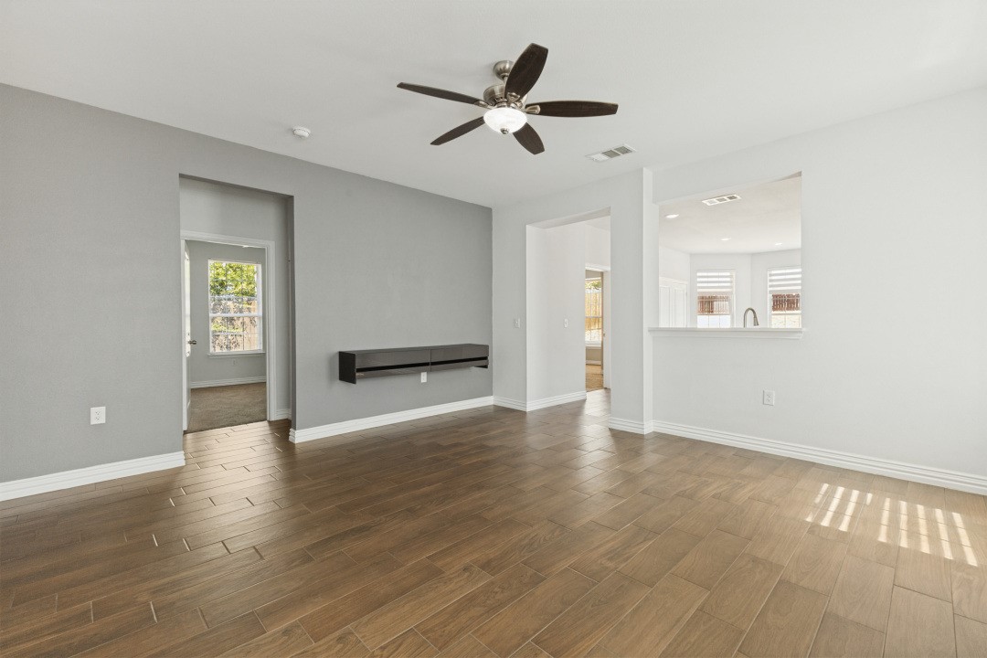 Empty living room with gray walls, wood-look tile flooring, and a ceiling fan beside an open kitchen pass-through.