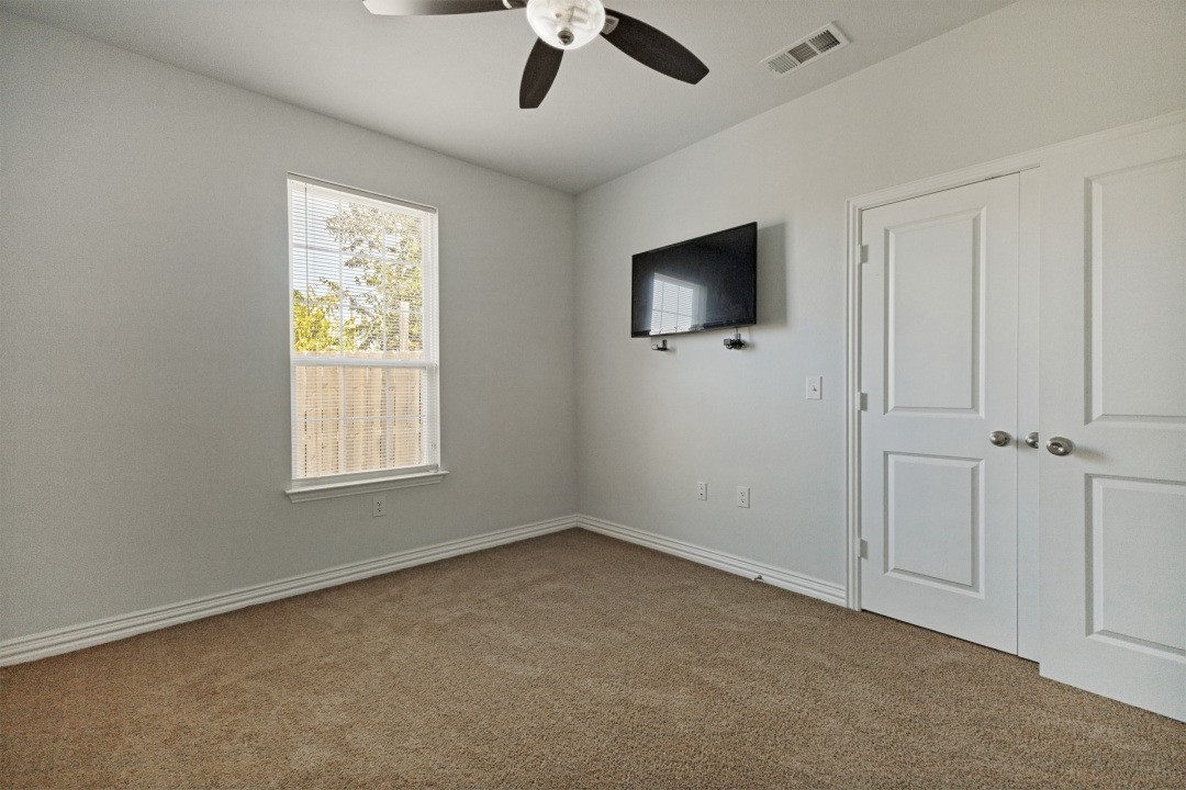 Empty carpeted bedroom with a wall-mounted television, window with blinds, and white closet doors.