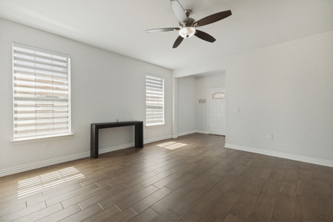 Mostly vacant living room with plank flooring, two shaded windows, a console table, and ceiling fan.