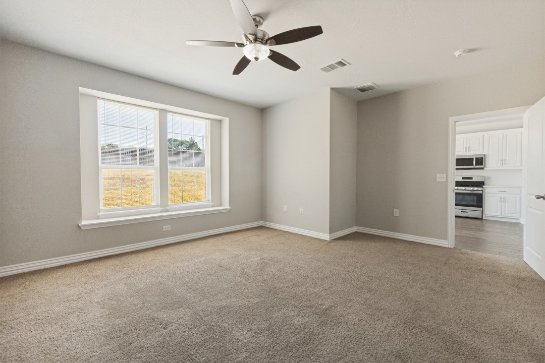 Empty carpeted room with a ceiling fan, wide window, and doorway to the kitchen.