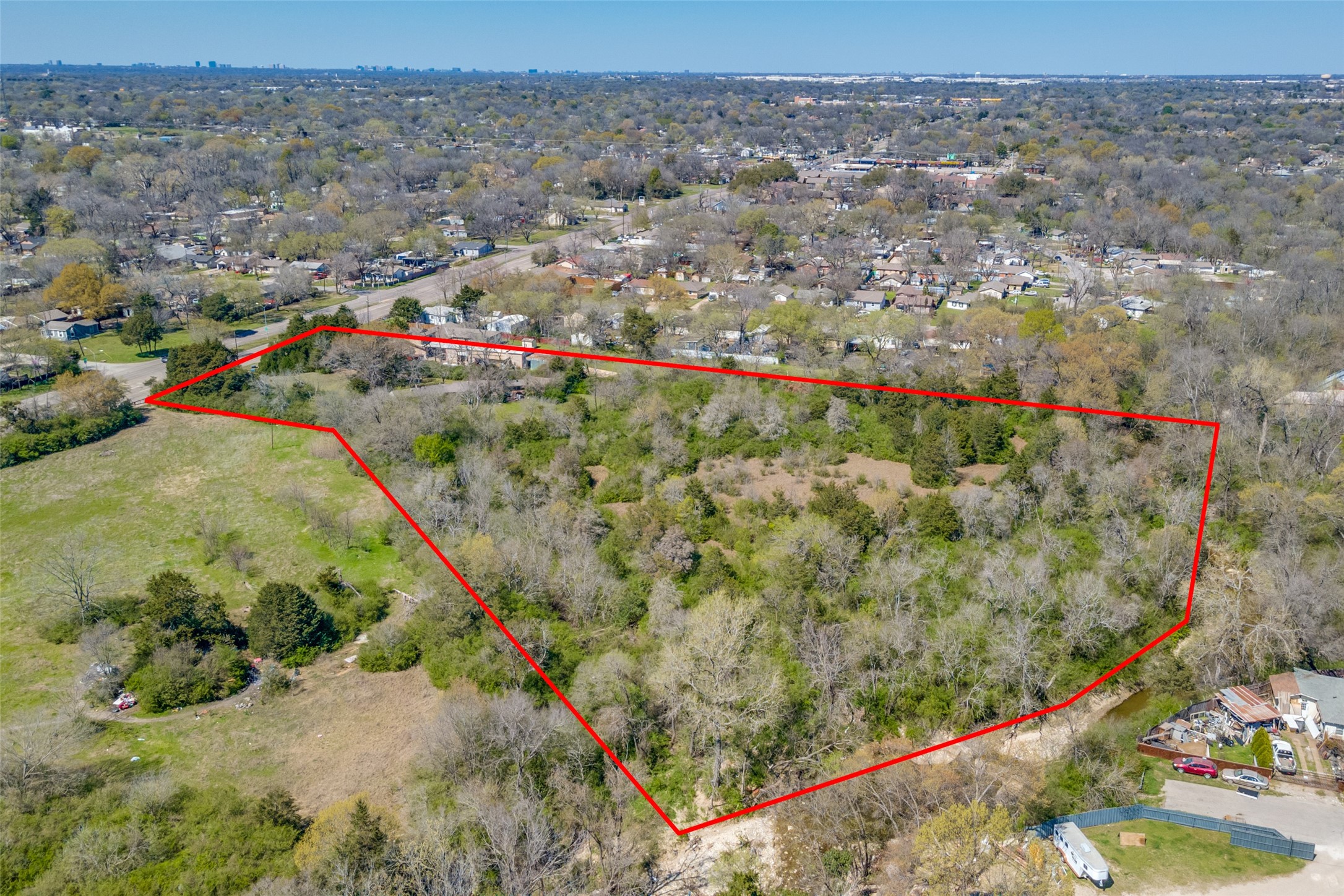 Aerial view of wooded vacant land outlined in red, with grassy areas and homes nearby.
