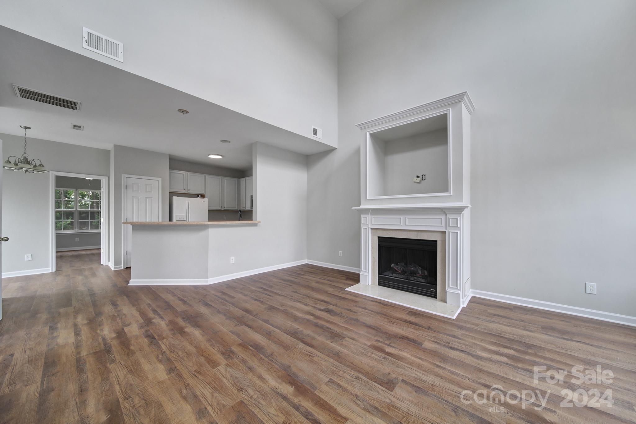 Empty living room with wood floors, a white fireplace, and an open kitchen with a breakfast bar.