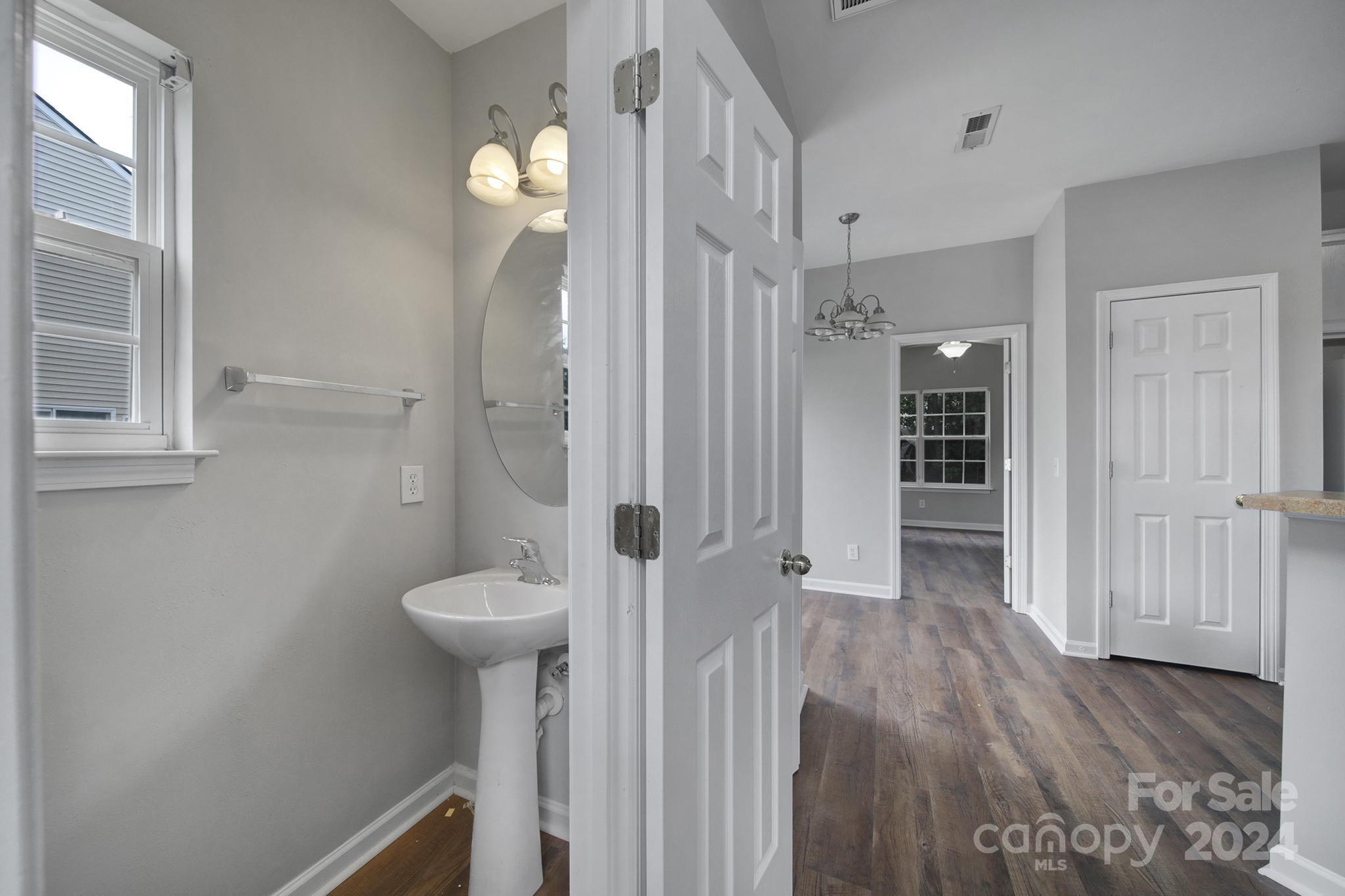 Bathroom with pedestal sink and oval mirror, looking through an open door into a wood-floored hallway.