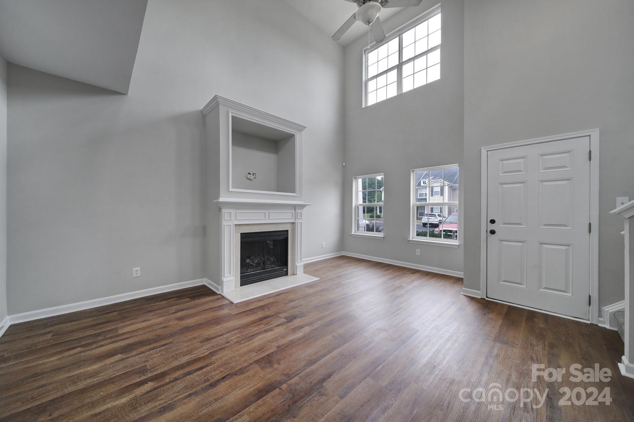 Vacant living room with a white fireplace, dark wood floors, tall windows, and a double front door.
