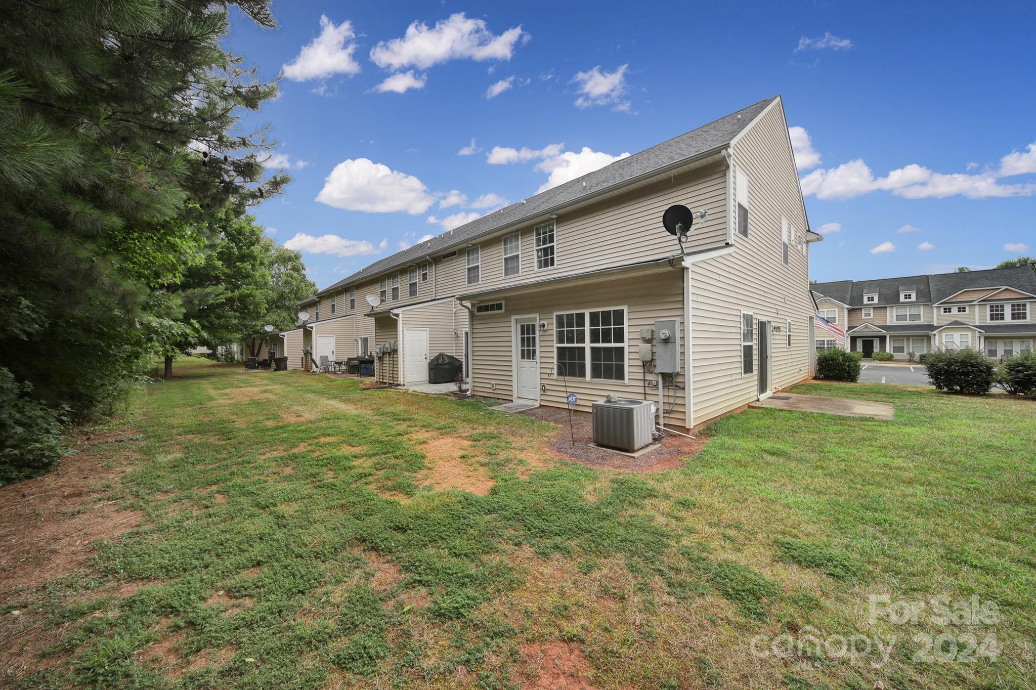 Rear of a beige two-story townhouse with a grassy yard, mature trees, and an outdoor air-conditioning unit.