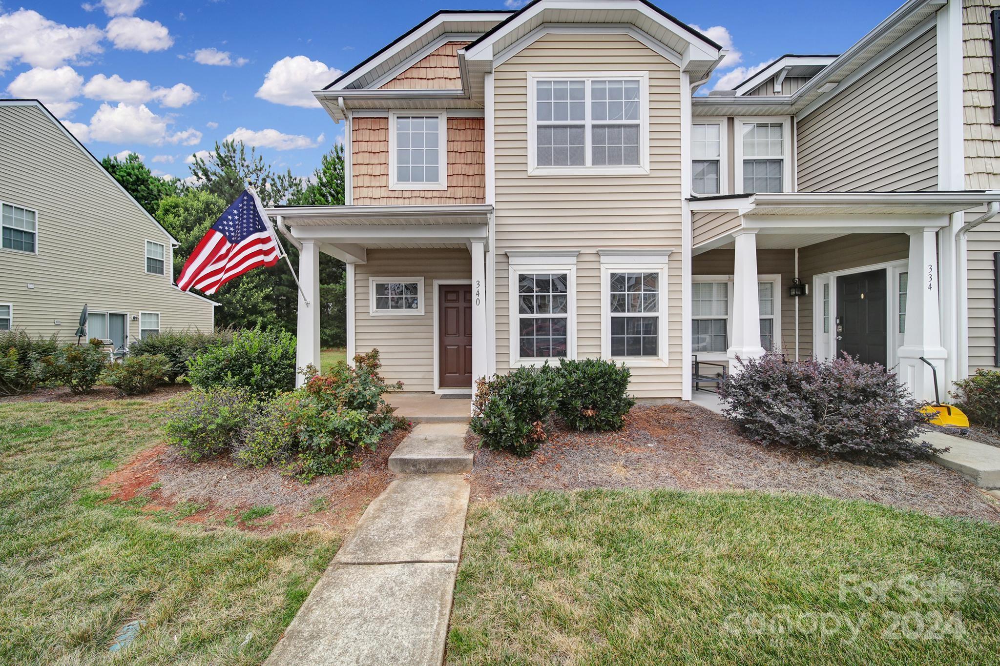 Two-story beige house with a covered porch, front walkway, landscaping, and an American flag.
