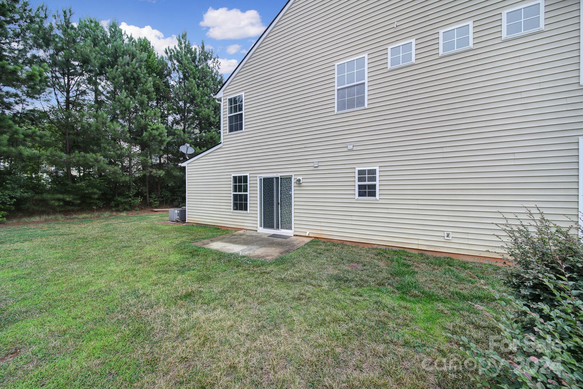 Rear of a beige two-story home with a sliding glass door opening onto a grassy yard bordered by pine trees.