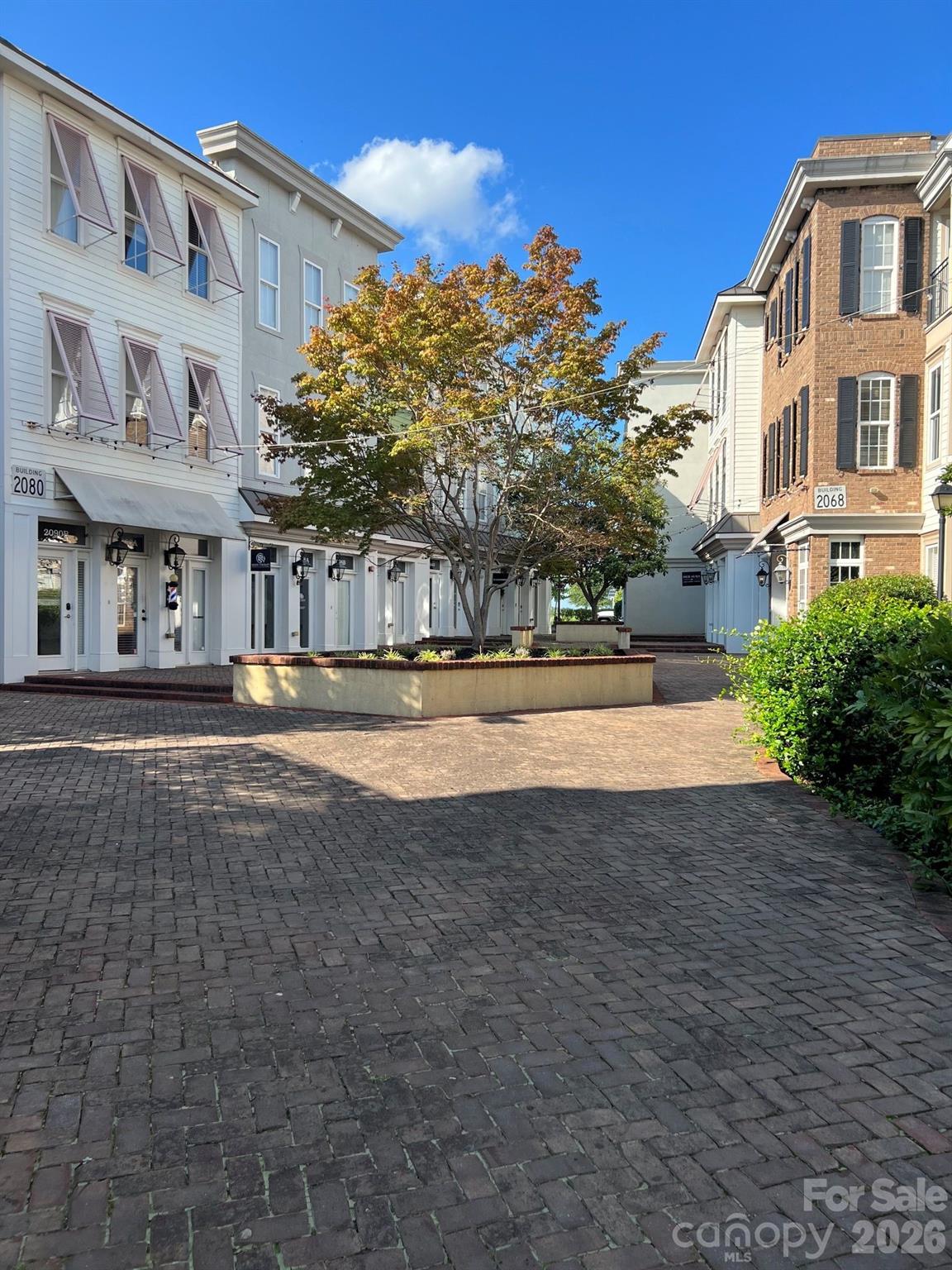 Commercial buildings frame a brick-paved courtyard with a central planter and autumn-colored tree.