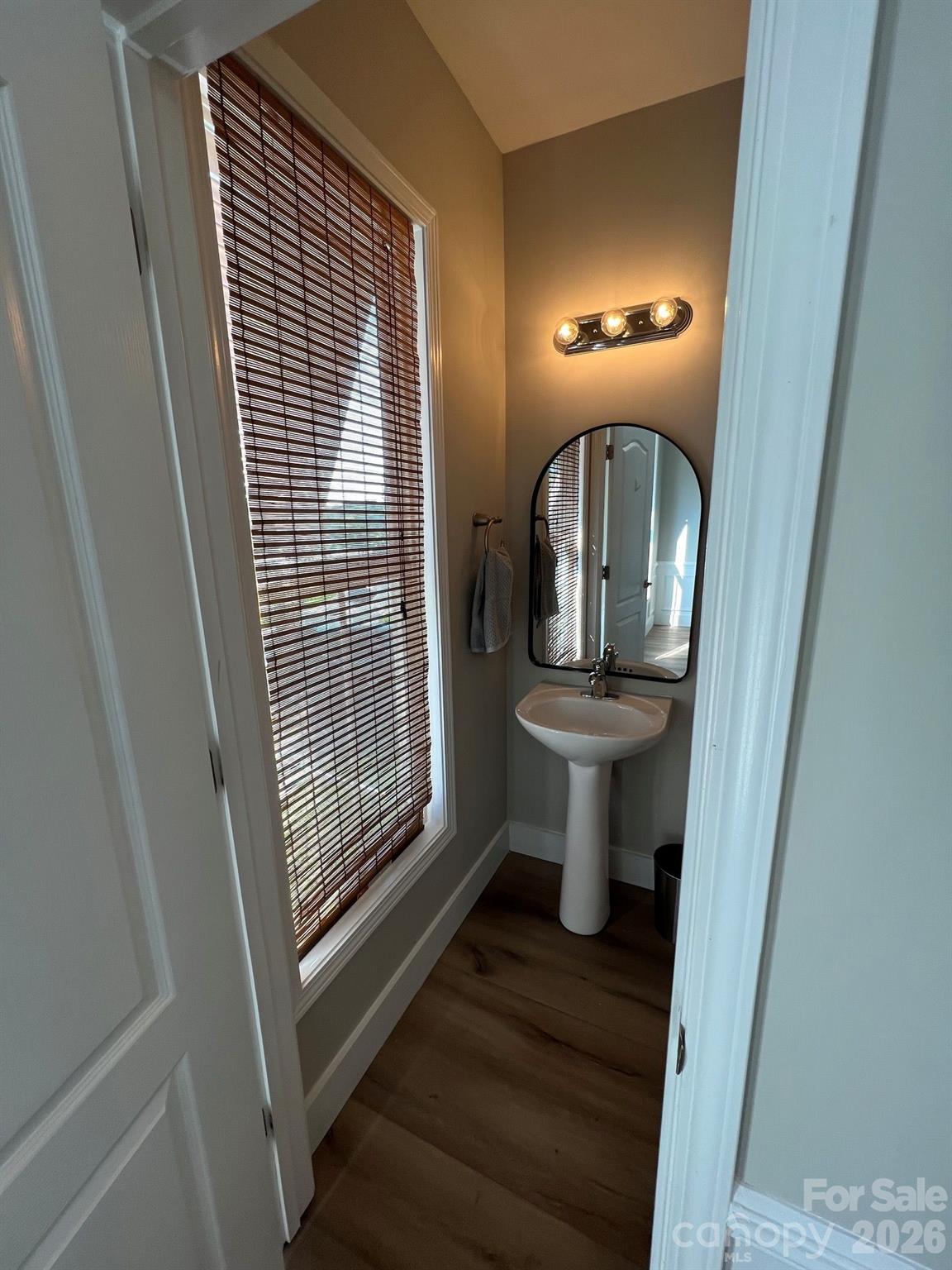 Powder room with a pedestal sink and arched mirror beside a tall window with wooden blinds.