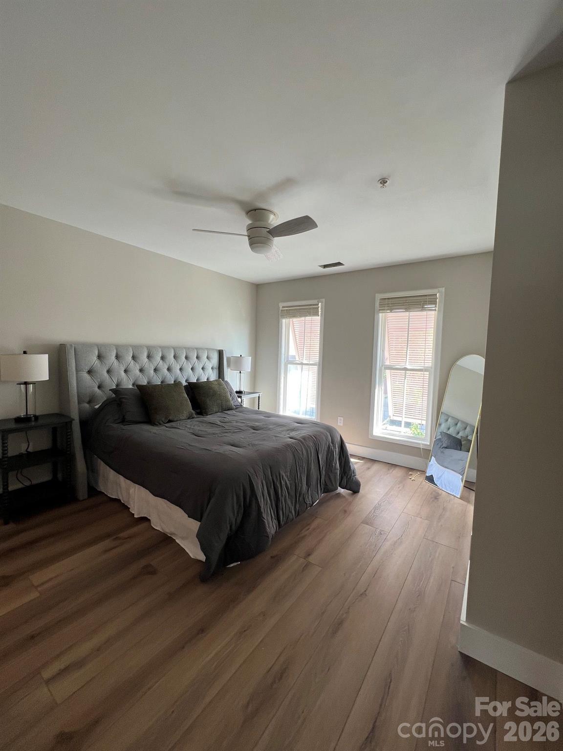 Furnished bedroom with a gray tufted bed, two windows, ceiling fan, and wood-plank flooring.