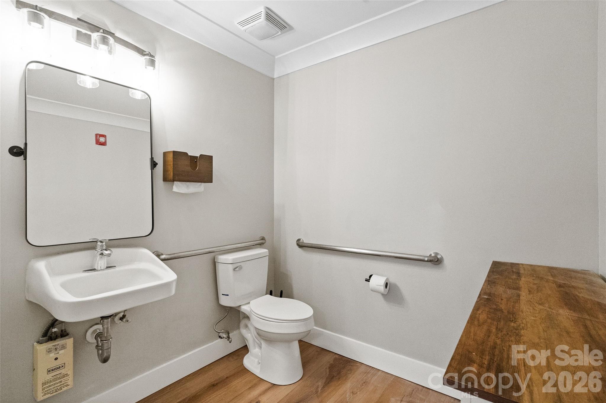 Bathroom with accessibility grab bars, wall-mounted sink, toilet, and wood-look flooring.