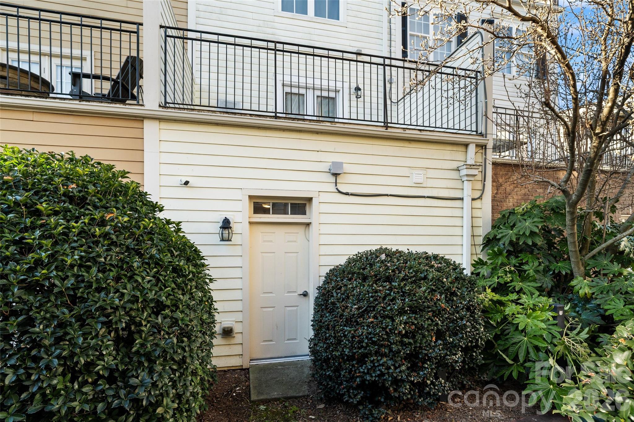 Home’s rear entry with a ground-level door, balcony railings, and surrounding shrubs.