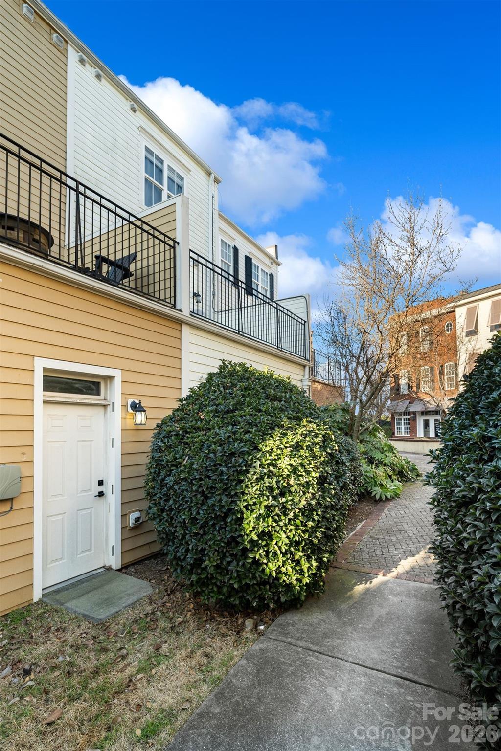 Townhouse exterior with black-railed balconies, a white side entrance, shrubs, and a brick walkway.