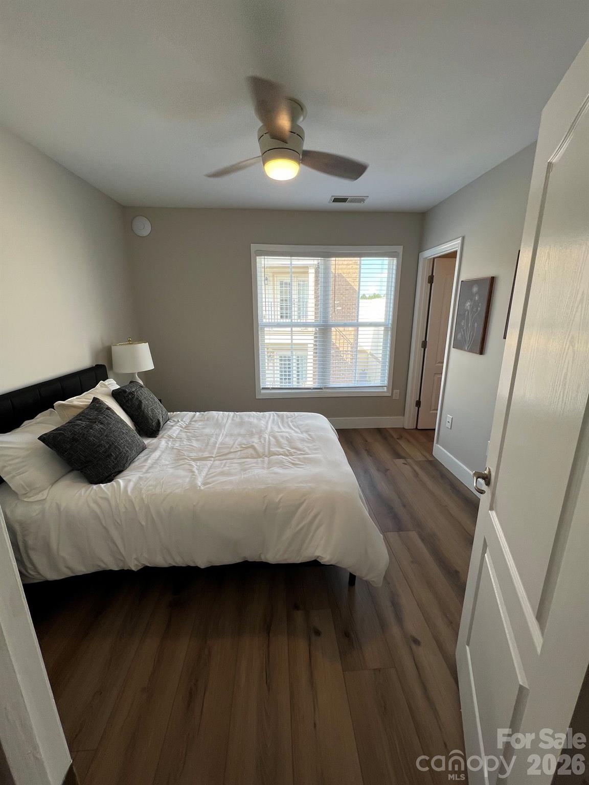 Furnished bedroom with a white bed, dark pillows, wood-look floors, and a ceiling fan.