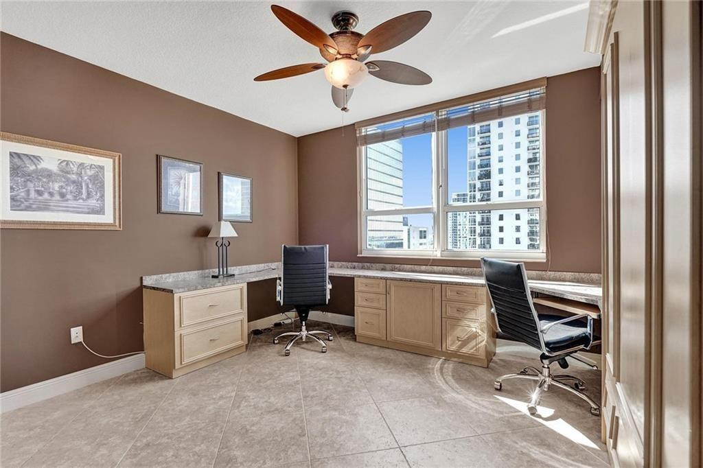 Home office with an L-shaped built-in desk, two chairs, and windows overlooking high-rise buildings.