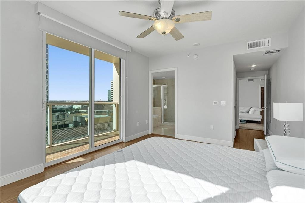 Bedroom with a white bed, ceiling fan, and sliding doors to a balcony overlooking the city.