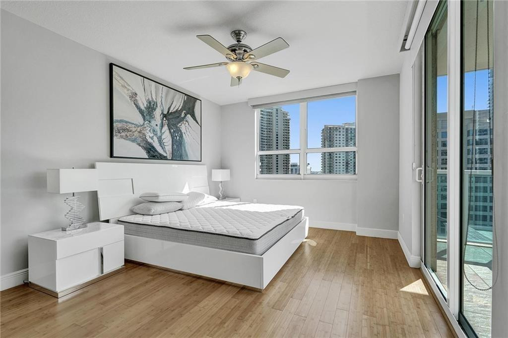 Condo bedroom with a white platform bed, abstract artwork, and balcony doors overlooking high-rise buildings.
