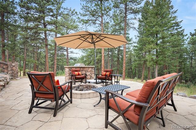 Stone patio with cushioned seating, a fire pit, and a large umbrella among pine trees.