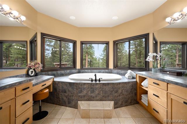 Bathroom with a built-in soaking tub, dual wood vanities, and large windows overlooking trees.
