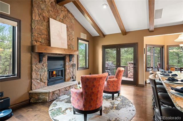 Living room with a tall stone fireplace, exposed ceiling beams, and wooded views through large windows.