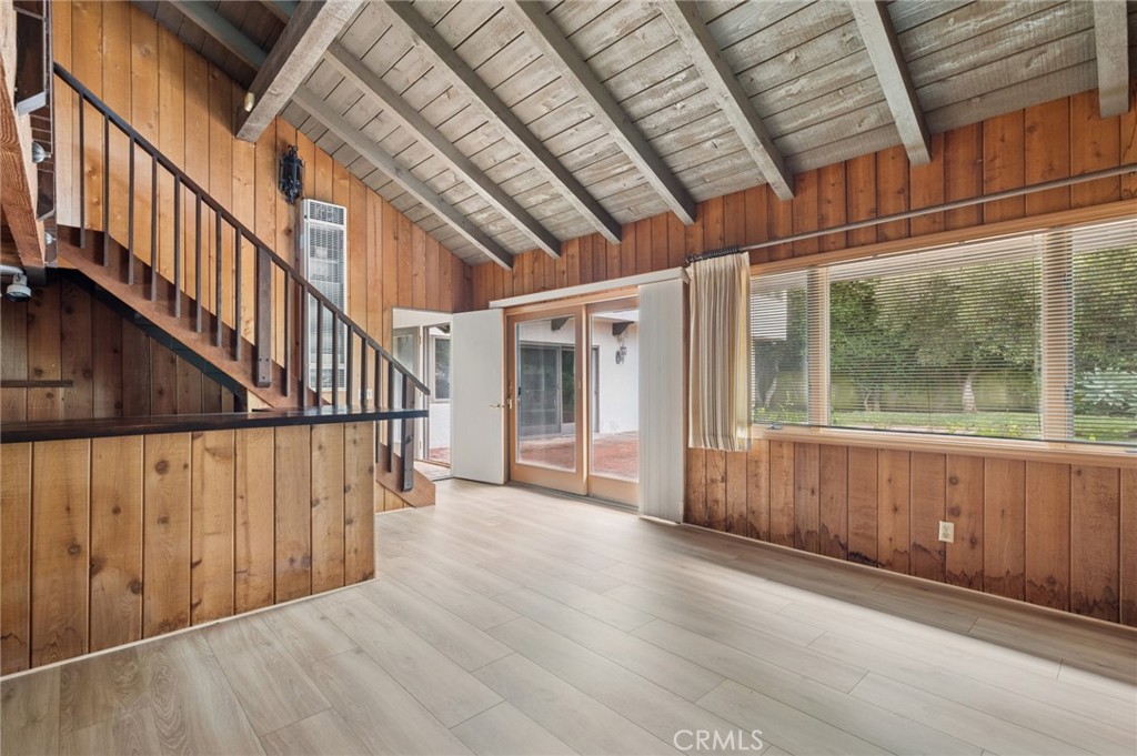 Empty living area with wood-paneled walls, vaulted beam ceiling, staircase, and large garden-facing window.