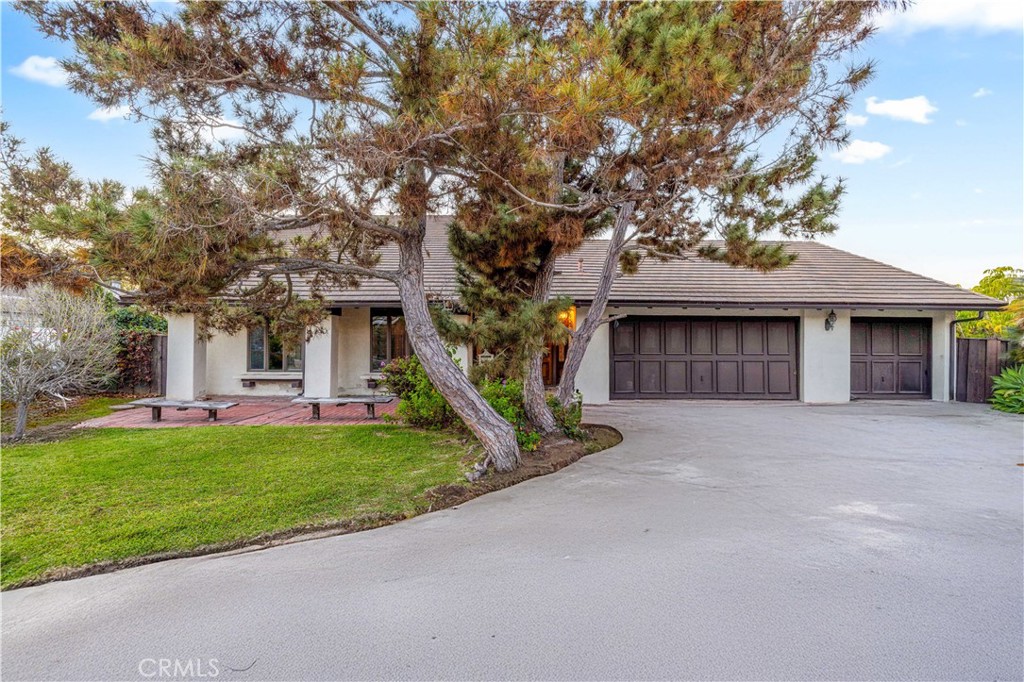 Single-story house with a broad driveway, two dark garage doors, and mature pine trees in front.