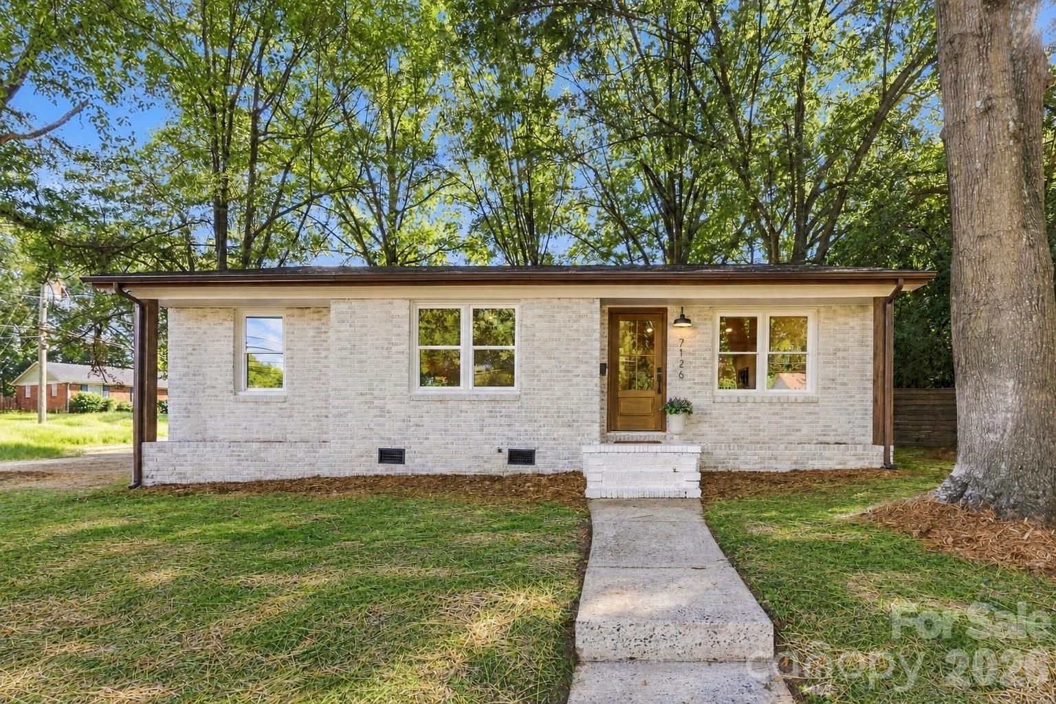White brick house with a wood front door, shaded by mature trees beside a concrete walkway.