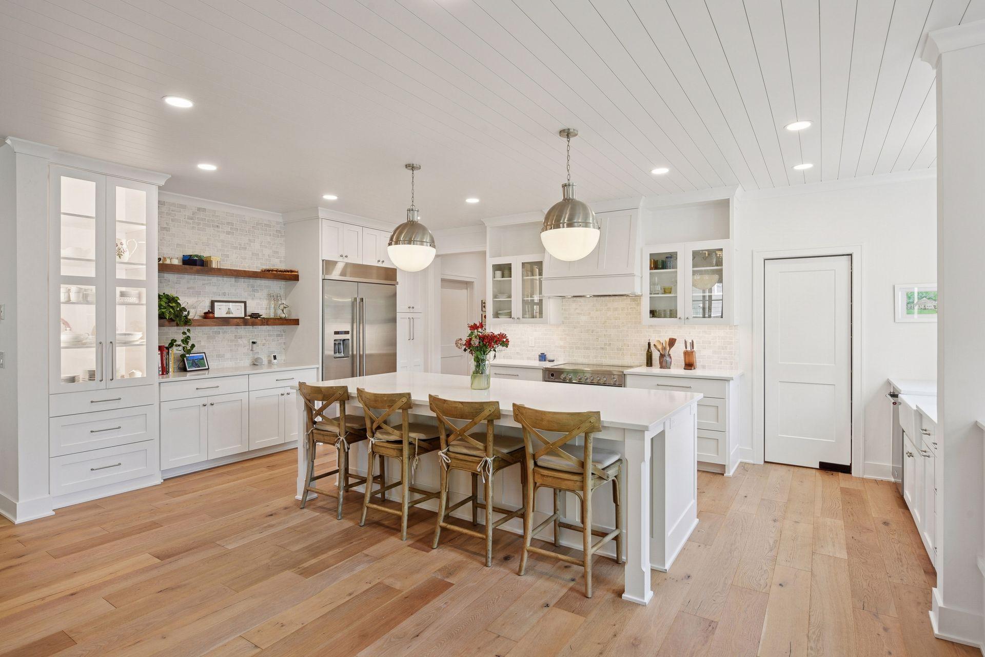 Bright white kitchen with a large island, wooden stools, glass-front cabinets, and wide-plank wood floors.