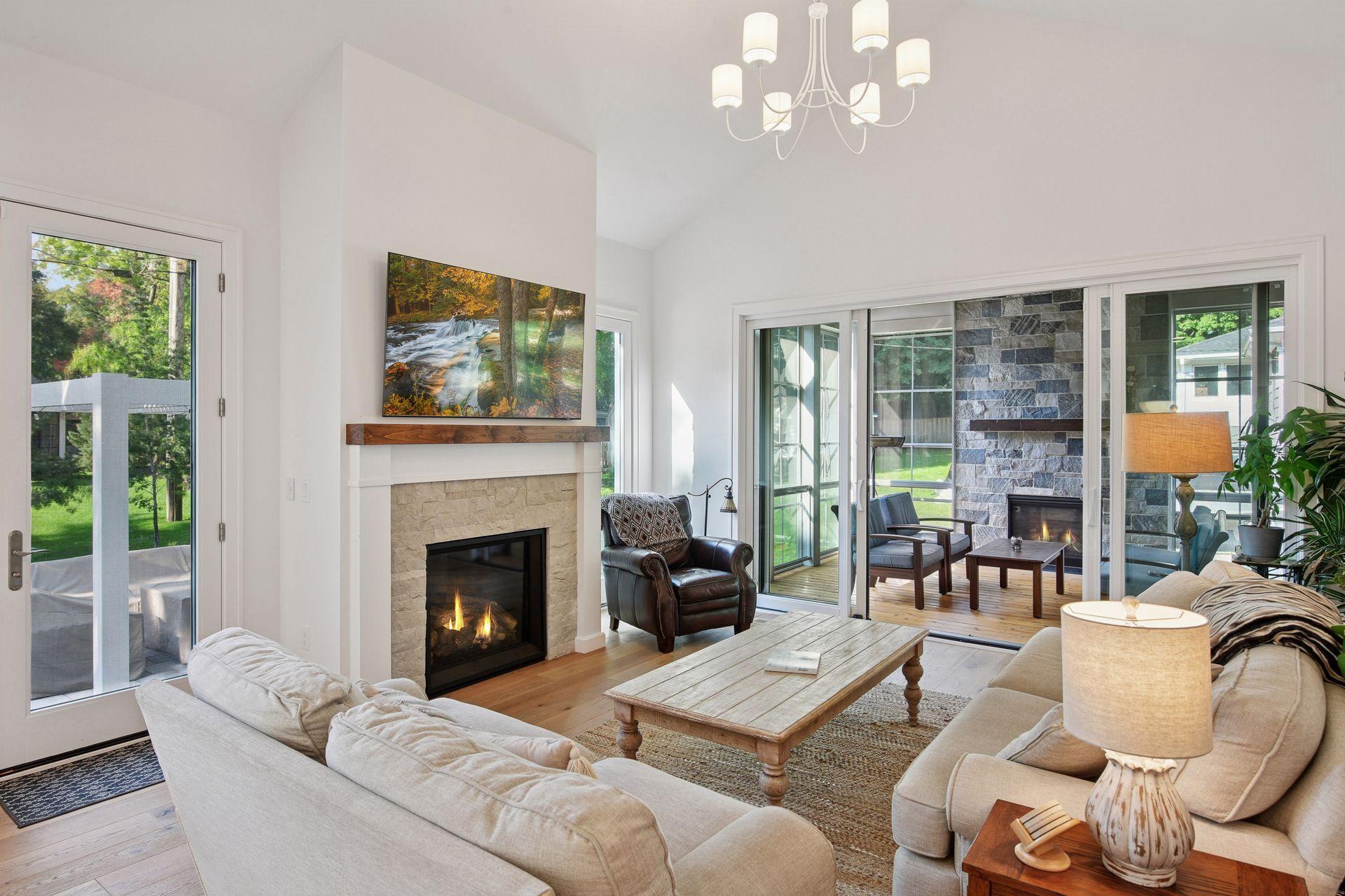 Living room with vaulted ceiling, stone fireplace, pale sofas, and glass doors to a patio fireplace.