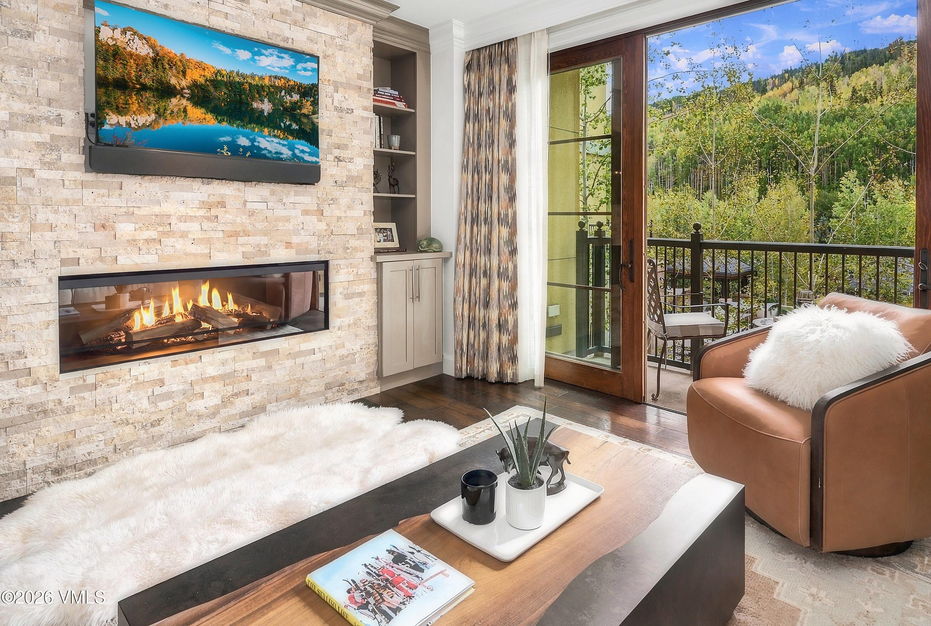 Living room with a stone fireplace and balcony doors overlooking autumn trees.