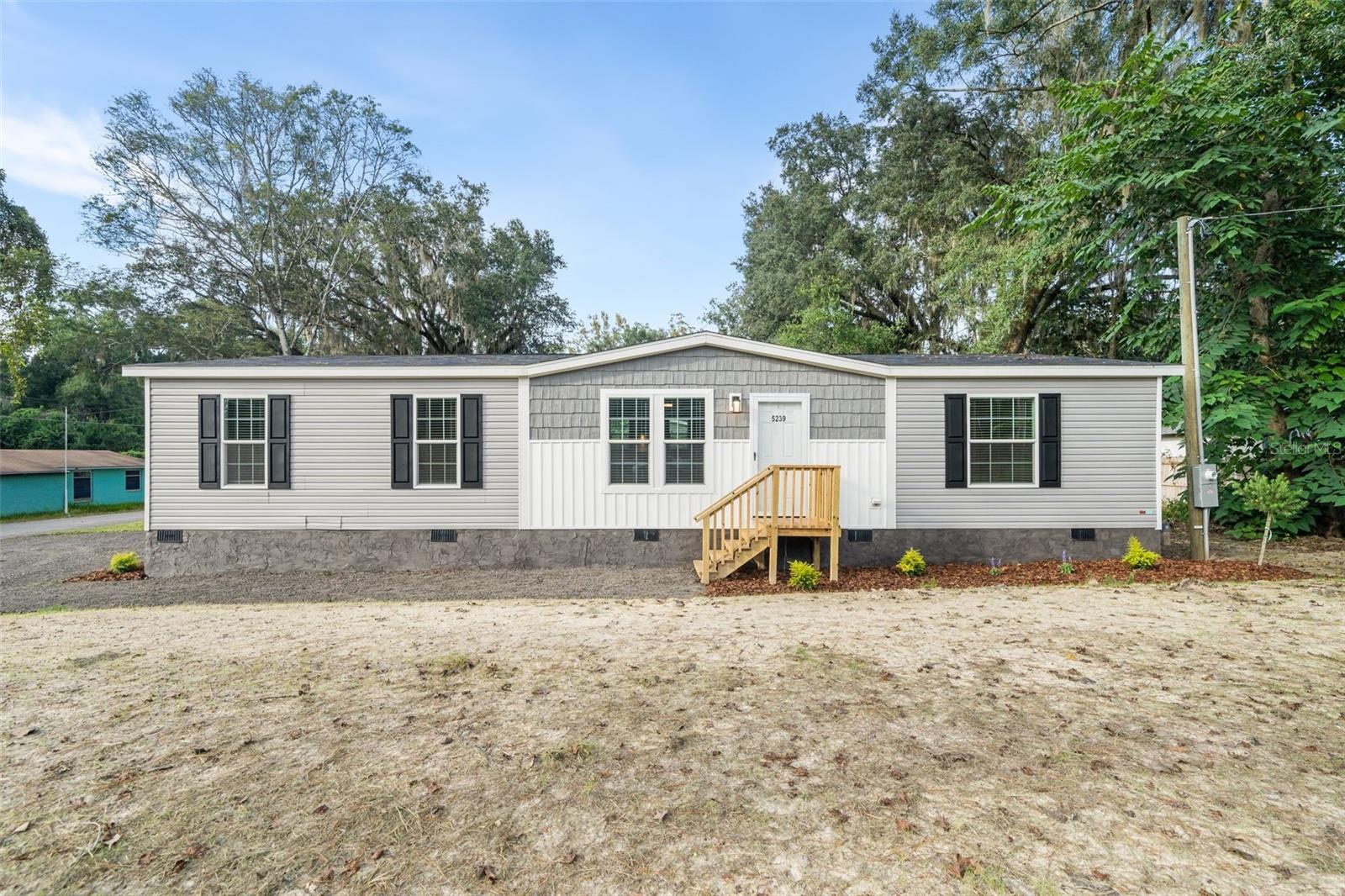 Gray manufactured home with central gable, dark shutters, and wooden front steps on a sandy lot.