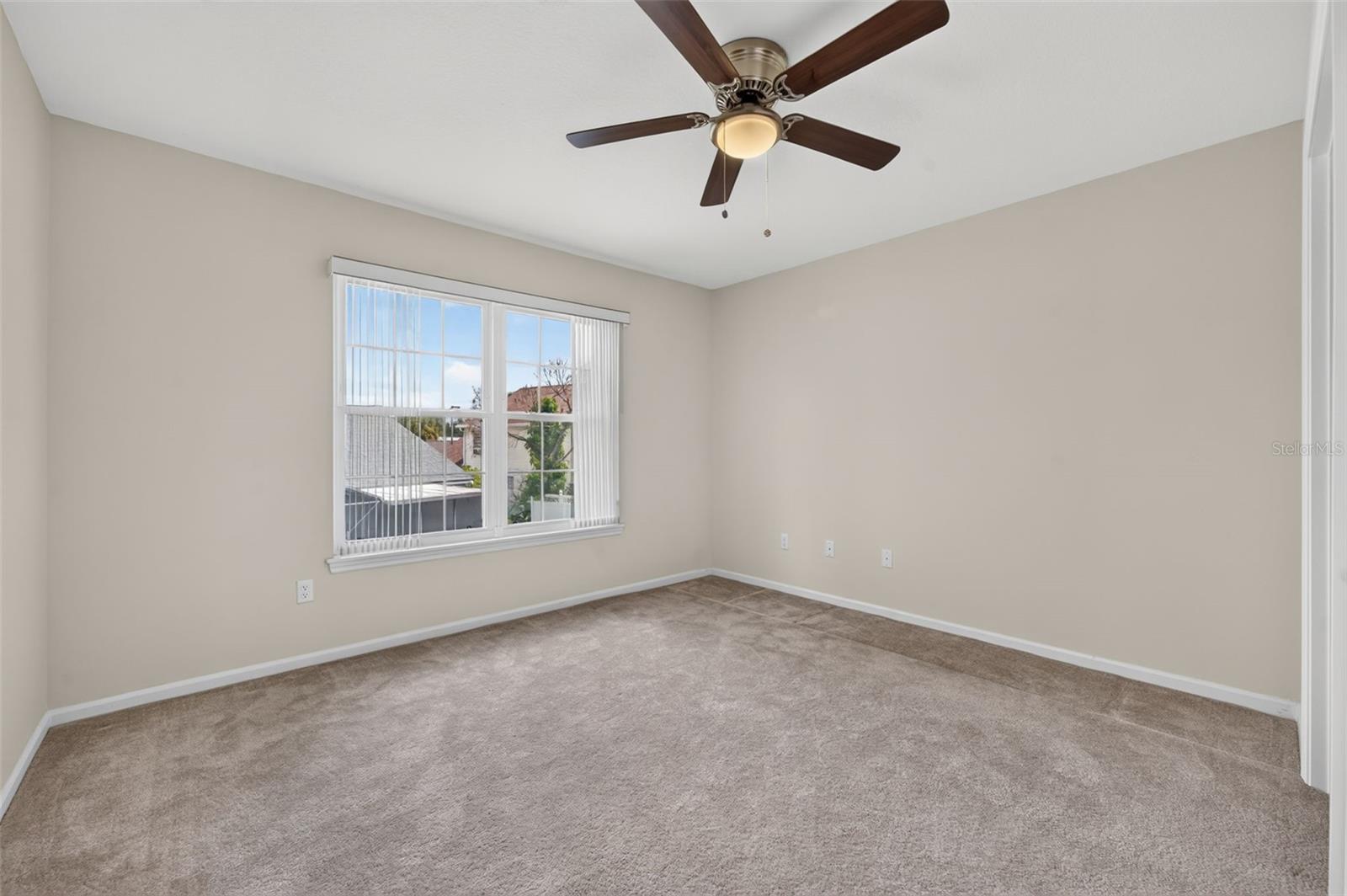 Empty carpeted bedroom with a ceiling fan and a window with vertical blinds.