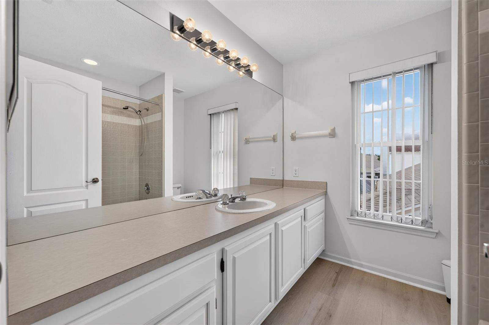 Bathroom with a long double-sink vanity, wall-length mirror, and beige tiled shower.