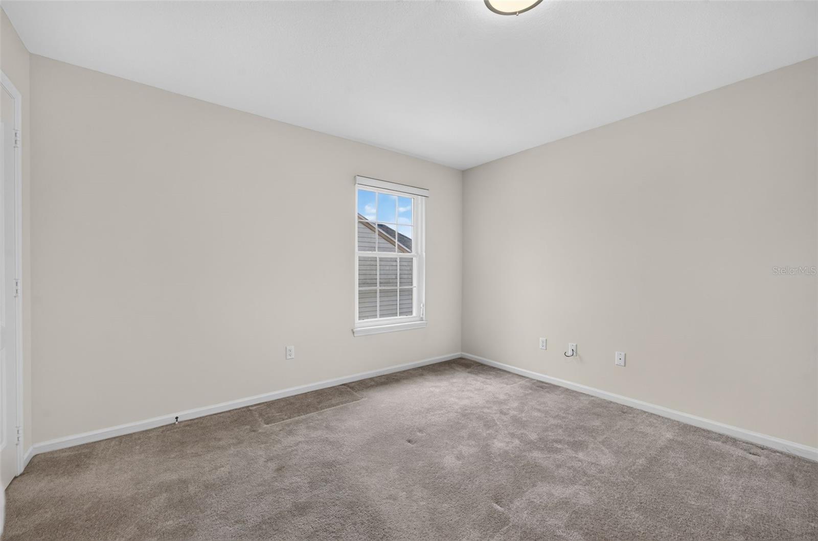 Empty bedroom with gray carpet, beige walls, and a window overlooking a neighboring roof.