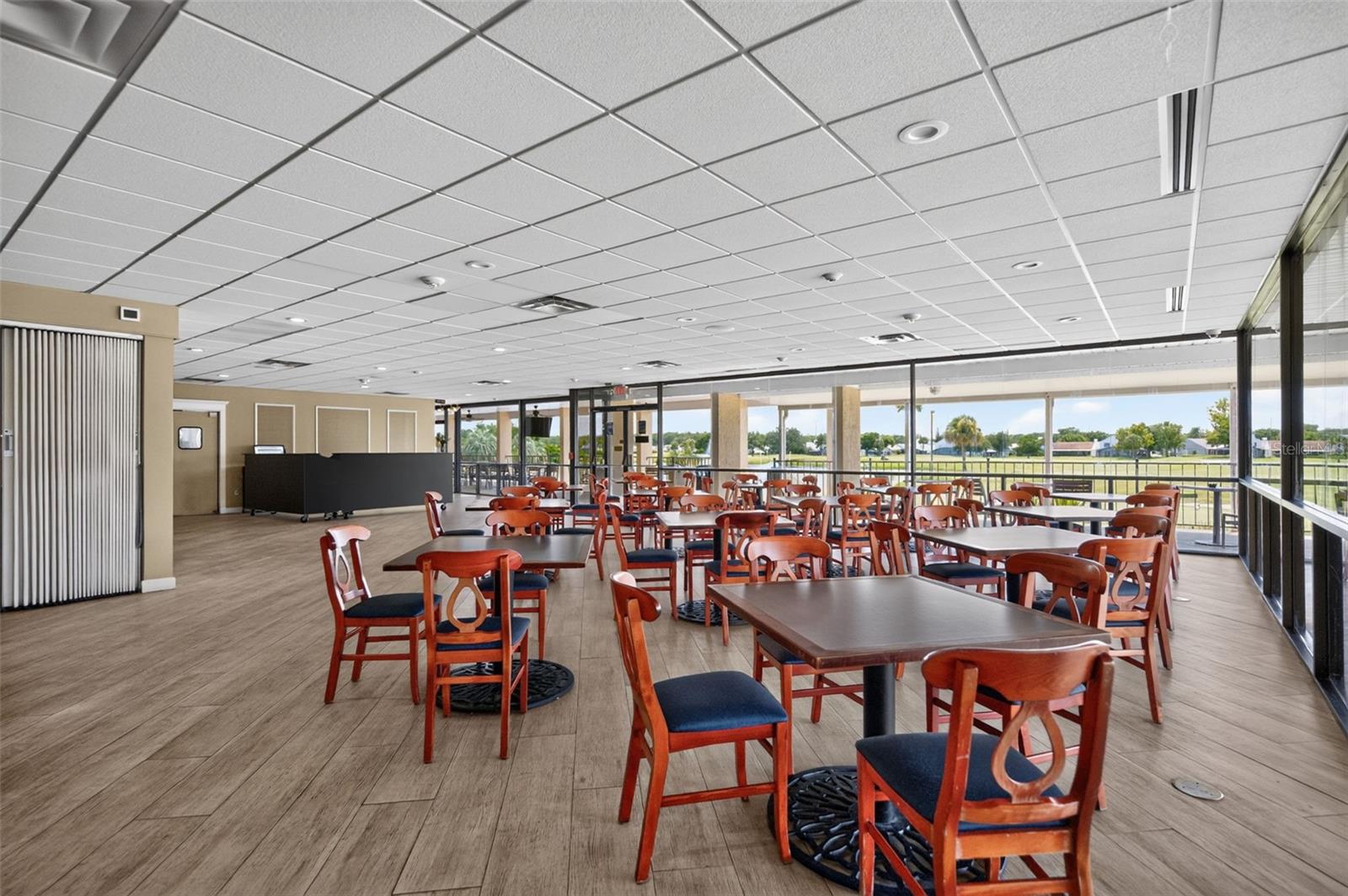 Community dining room with rows of wooden tables and chairs beside windows overlooking green grounds.