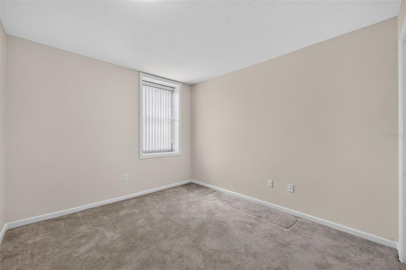Empty carpeted room with beige walls and a narrow window with vertical blinds.