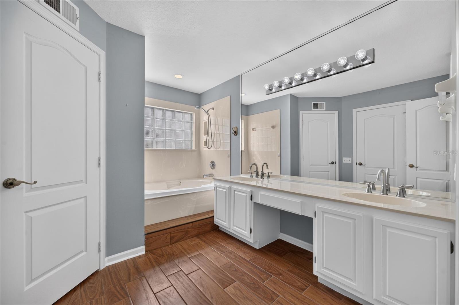 Bathroom with a double-sink vanity, large mirror, bathtub, and glass-block window.
