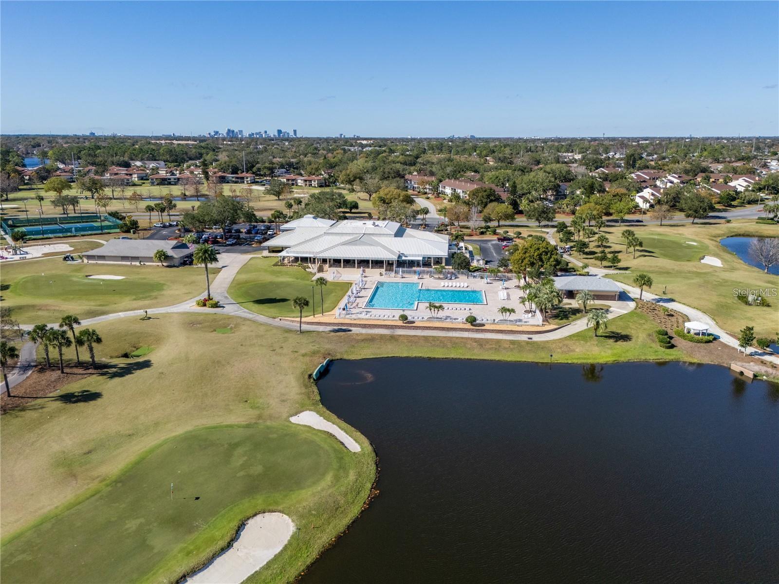Aerial view of a golf course clubhouse and pool beside a pond, with Orlando's skyline in the distance.