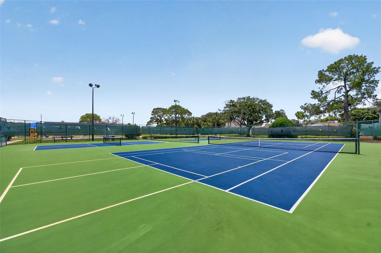 Outdoor tennis courts with blue playing areas, green surrounds, and perimeter fencing.