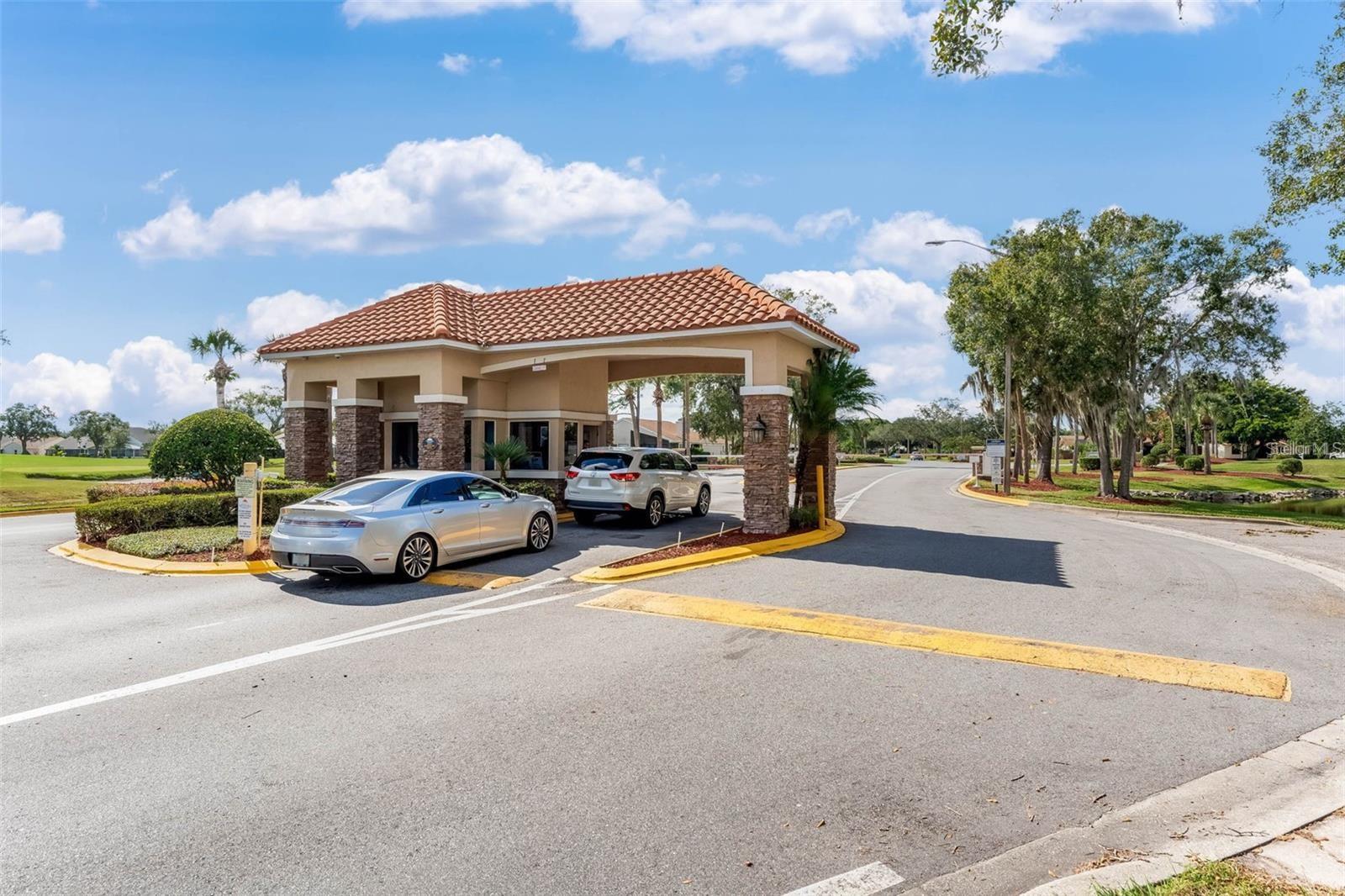Community entrance pavilion with a terracotta roof and stone columns beside a landscaped drive.