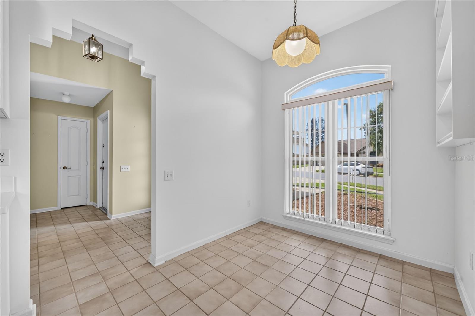 Empty tiled dining area with an arched window and vertical blinds beside a pale yellow hallway.
